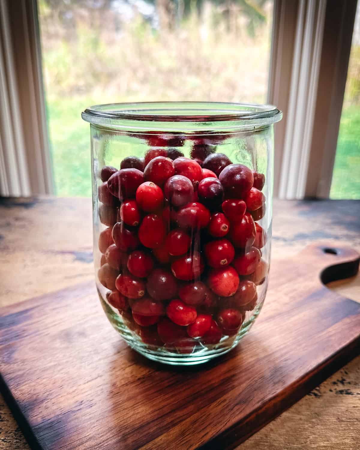 Fresh cranberries in a jar that is resting on a wood cutting board with light shining through a window
